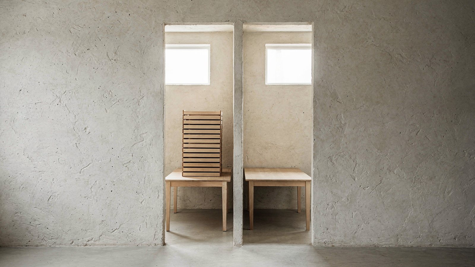 Two identical bare wooden tables in two identical rooms, seen through a split opening in a plaster wall. The left table carries a tall stack of nested wooden trays; the right table is empty.